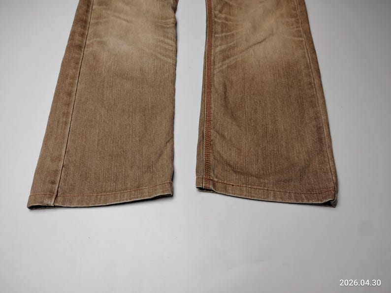 May include: Close-up of the lower portion of a pair of brown denim jeans. The jeans have a faded appearance and visible stitching along the seams. The fabric appears to be a medium weight denim.