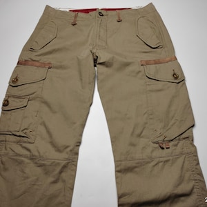 May include: Khaki-colored cargo pants with multiple pockets and brown leather accents. The pants have a button and zipper closure, and a relaxed fit. The image shows the pants laid flat on a white surface.
