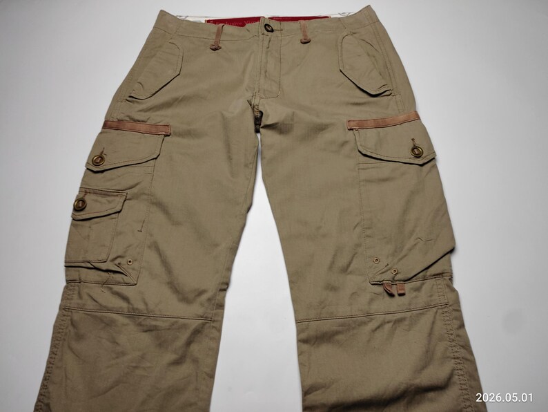 May include: Khaki-colored cargo pants with multiple pockets and brown leather accents. The pants have a button and zipper closure, and a relaxed fit. The image shows the pants laid flat on a white surface.