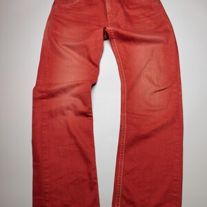 May include: Red denim jeans with a classic five-pocket design. The jeans have a button closure and white stitching along the seams. The fabric has a faded appearance, giving it a vintage look.