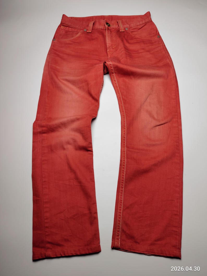 May include: Red denim jeans with a classic five-pocket design. The jeans have a button closure and white stitching along the seams. The fabric has a faded appearance, giving it a vintage look.
