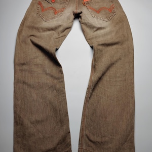 May include: Brown denim jeans with orange stitching and zipper accents. Features include two back pockets with orange stitching, a small pocket on the right side, and a zipper closure on the back right pocket. The jeans have a straight leg cut.