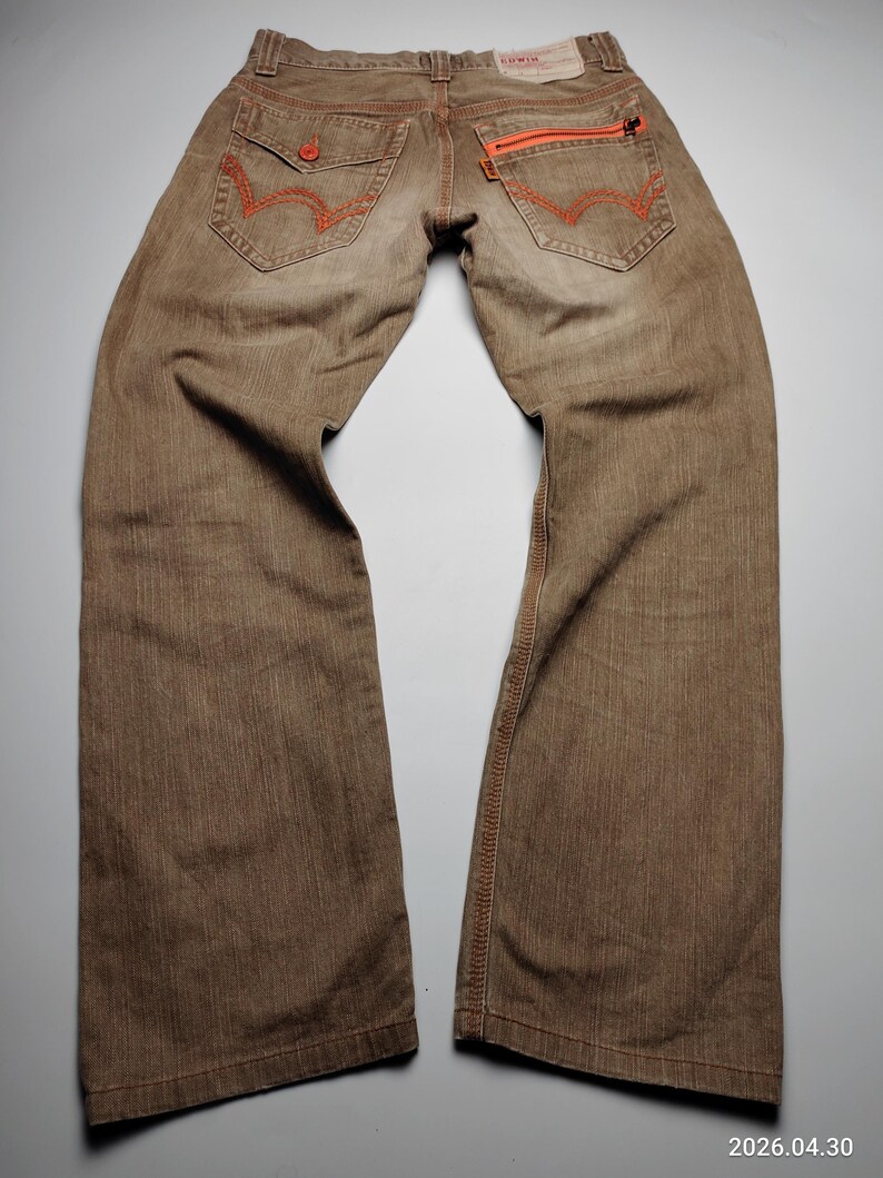 May include: Brown denim jeans with orange stitching and zipper accents. Features include two back pockets with orange stitching, a small pocket on the right side, and a zipper closure on the back right pocket. The jeans have a straight leg cut.