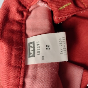 May include: Close-up of red denim fabric with a white tag. The tag displays the brand "Edwin", the style "483XVS", and the size "30". Additional codes are also printed on the tag.