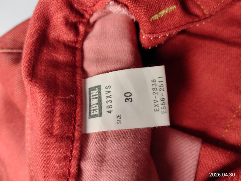 May include: Close-up of red denim fabric with a white tag. The tag displays the brand "Edwin", the style "483XVS", and the size "30". Additional codes are also printed on the tag.