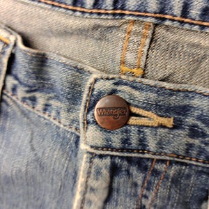 May include: Close-up of a pair of blue denim jeans. The jeans feature a copper-colored button with the brand name "Wrangler" embossed on it. The stitching is visible, and the fabric shows signs of wear.