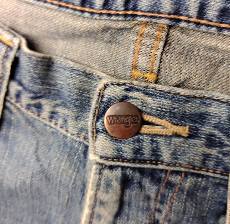 May include: Close-up of a pair of blue denim jeans. The jeans feature a copper-colored button with the brand name "Wrangler" embossed on it. The stitching is visible, and the fabric shows signs of wear.