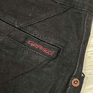 Puede incluir: Primer plano de unos pantalones vaqueros negros con un bolsillo visible y detalles de costura. El logotipo rojo "GRAMICCI" est&aacute; bordado en el bolsillo. Se ve un bot&oacute;n color cobre en el lateral.