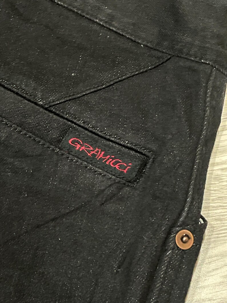 Puede incluir: Primer plano de unos pantalones vaqueros negros con un bolsillo visible y detalles de costura. El logotipo rojo "GRAMICCI" est&aacute; bordado en el bolsillo. Se ve un bot&oacute;n color cobre en el lateral.