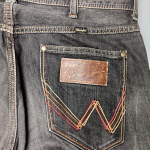 May include: Close-up of black denim jeans with a brown leather patch on the back pocket. The patch is embossed with the Wrangler logo. The pocket features a decorative W-shaped stitching in red, orange, and yellow.