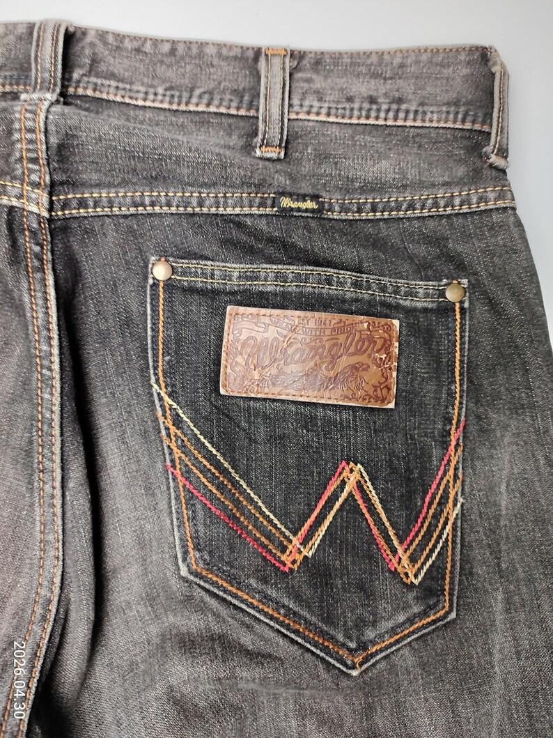 May include: Close-up of black denim jeans with a brown leather patch on the back pocket. The patch is embossed with the Wrangler logo. The pocket features a decorative W-shaped stitching in red, orange, and yellow.