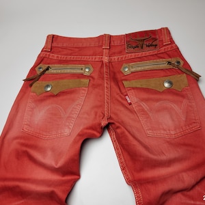 May include: Red denim jeans with brown suede pocket accents and zipper details. The jeans feature a leather patch with embossed text and a small red tag. The back pockets have a classic design.