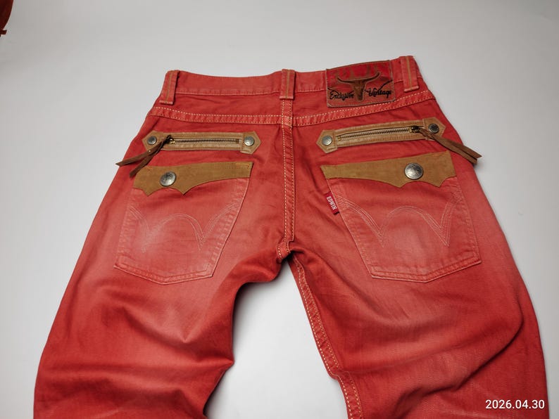 May include: Red denim jeans with brown suede pocket accents and zipper details. The jeans feature a leather patch with embossed text and a small red tag. The back pockets have a classic design.