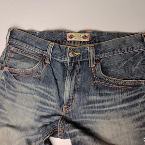 May include: Close-up of blue denim jeans with a faded wash. The jeans feature classic five-pocket styling, with orange stitching details around the pockets and seams. A small, patterned tag is visible on the waistband.