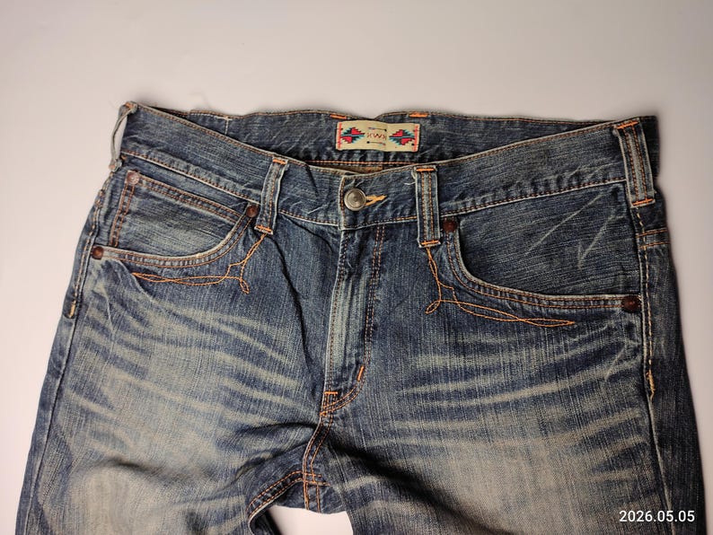 May include: Close-up of blue denim jeans with a faded wash. The jeans feature classic five-pocket styling, with orange stitching details around the pockets and seams. A small, patterned tag is visible on the waistband.