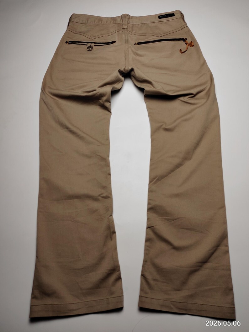 May include: Tan-colored pants with a classic five-pocket design. The back pockets feature black zippers and decorative stitching. The pants are made of a durable fabric and have a straight leg cut.