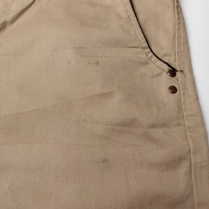May include: Close-up of khaki-colored pants. The pants feature a front pocket with two decorative rivets. The fabric has a subtle texture and visible stitching. The pants are made of a durable material, suitable for everyday wear.