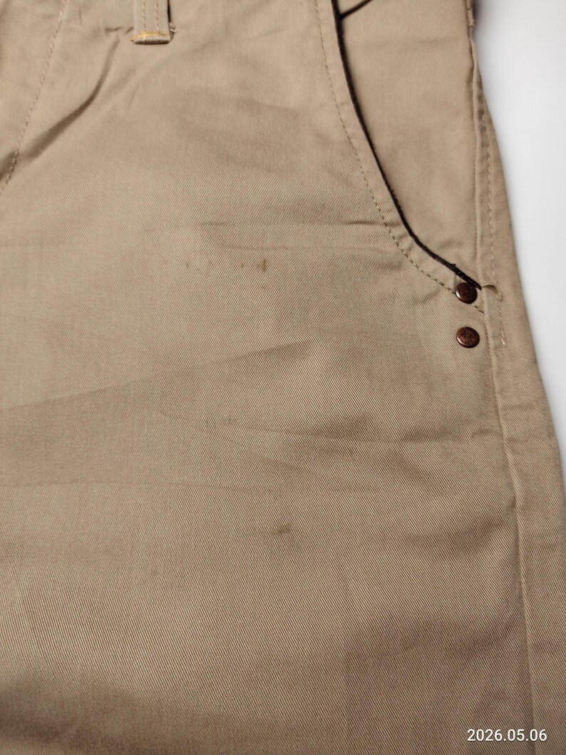 May include: Close-up of khaki-colored pants. The pants feature a front pocket with two decorative rivets. The fabric has a subtle texture and visible stitching. The pants are made of a durable material, suitable for everyday wear.