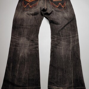 May include: Dark wash denim jeans with a flared leg. The back pockets feature a brown leather patch with orange and red stitching. The jeans have a worn look and a classic Western style.