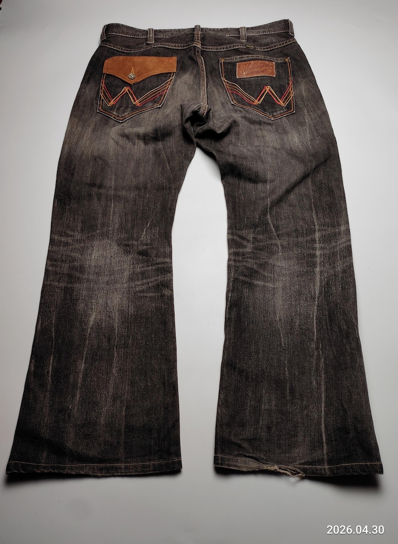 May include: Dark wash denim jeans with a flared leg. The back pockets feature a brown leather patch with orange and red stitching. The jeans have a worn look and a classic Western style.