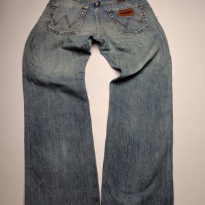 May include: Faded blue denim jeans with a classic five-pocket design. The jeans feature the Wrangler brand label on the back pocket and a worn appearance. The jeans are laid flat, showcasing the back view.