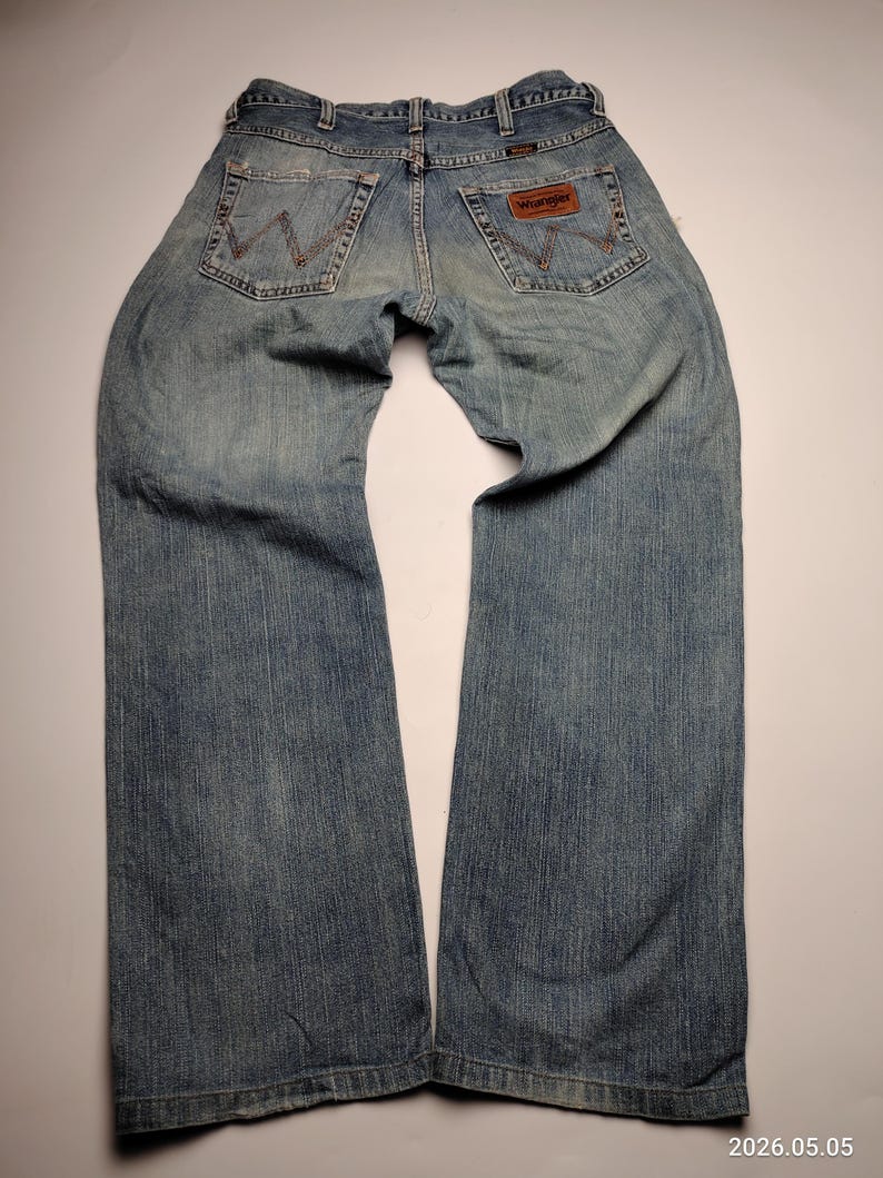 May include: Faded blue denim jeans with a classic five-pocket design. The jeans feature the Wrangler brand label on the back pocket and a worn appearance. The jeans are laid flat, showcasing the back view.