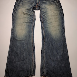 May include: Blue denim jeans with a faded wash and distressed details. The jeans have a classic five-pocket design, a button and zipper closure, and orange stitching. The hem is frayed. The jeans are laid flat on a white surface.