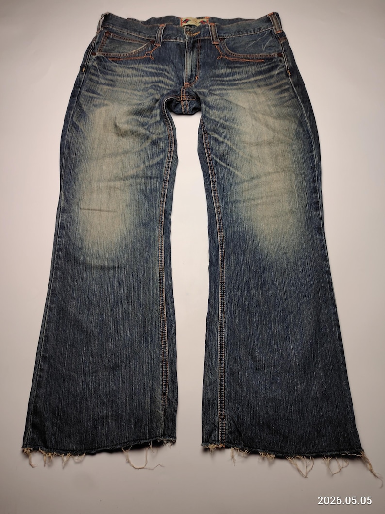 May include: Blue denim jeans with a faded wash and distressed details. The jeans have a classic five-pocket design, a button and zipper closure, and orange stitching. The hem is frayed. The jeans are laid flat on a white surface.