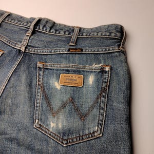 Peut inclure: Gros plan du dos d'un jean Wrangler en denim bleu. Le jean a un aspect us&eacute; avec des zones d&eacute;lav&eacute;es et un patch en cuir marron sur la poche arri&egrave;re. Le logo Wrangler est visible sur le patch et la ceinture.