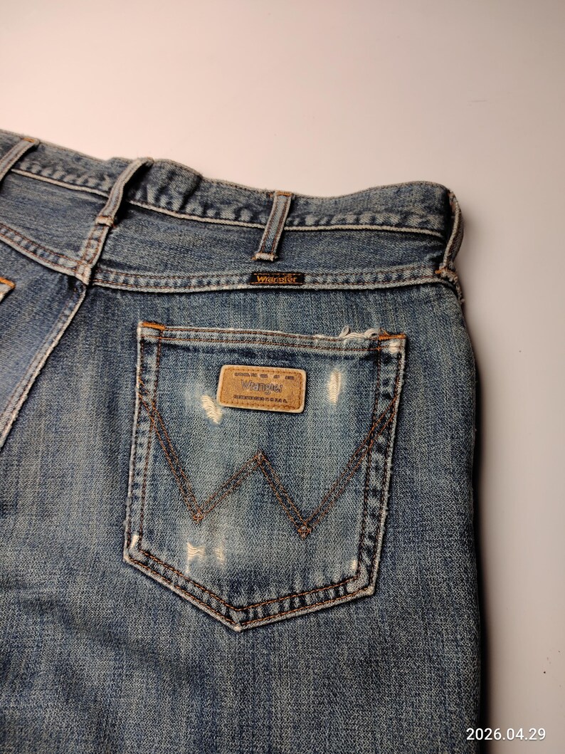 Peut inclure: Gros plan du dos d'un jean Wrangler en denim bleu. Le jean a un aspect us&eacute; avec des zones d&eacute;lav&eacute;es et un patch en cuir marron sur la poche arri&egrave;re. Le logo Wrangler est visible sur le patch et la ceinture.