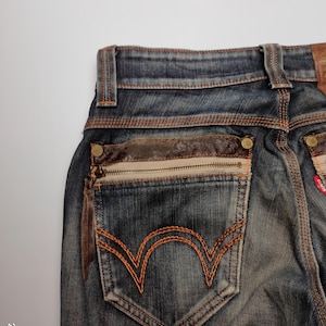 Puede incluir: Primer plano de la parte trasera de unos vaqueros azules con detalles en cuero marr&oacute;n. Los vaqueros tienen un bolsillo con cremallera y tirador de cuero y un dise&ntilde;o cosido en naranja en el bolsillo trasero.