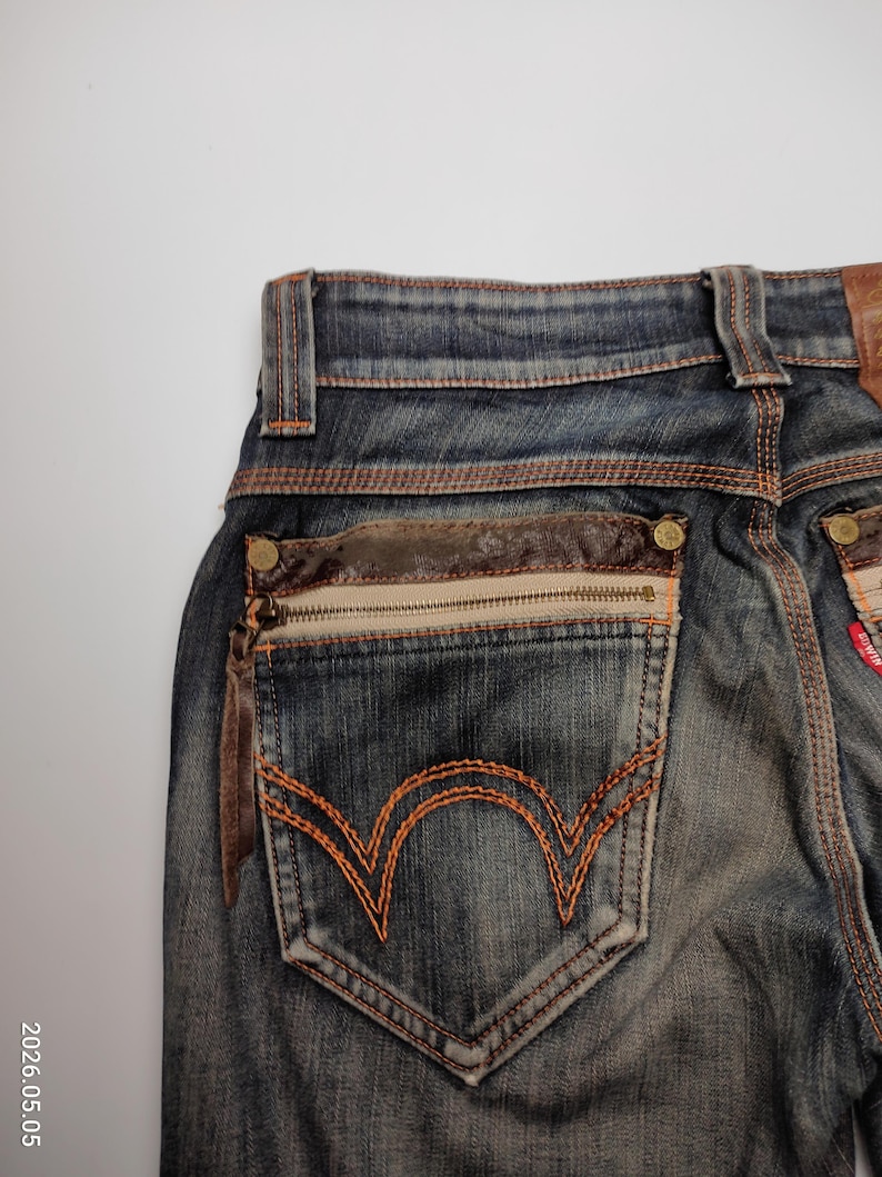 Puede incluir: Primer plano de la parte trasera de unos vaqueros azules con detalles en cuero marr&oacute;n. Los vaqueros tienen un bolsillo con cremallera y tirador de cuero y un dise&ntilde;o cosido en naranja en el bolsillo trasero.