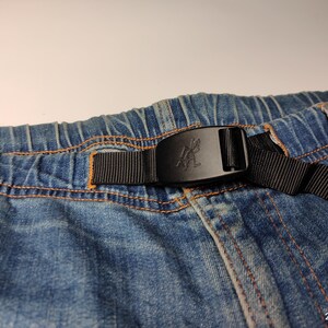 May include: Close-up of blue denim shorts with an elasticated waistband and a black belt. The belt has a black buckle with a logo. The shorts have orange stitching.