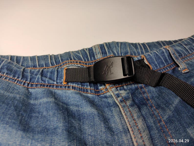 May include: Close-up of blue denim shorts with an elasticated waistband and a black belt. The belt has a black buckle with a logo. The shorts have orange stitching.
