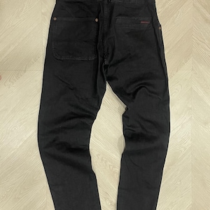 Puede incluir: Pantalones vaqueros negros con un dise&ntilde;o cl&aacute;sico de cinco bolsillos. Los pantalones tienen cierre de bot&oacute;n, trabillas para el cintur&oacute;n y una peque&ntilde;a etiqueta roja con el logotipo en el bolsillo trasero. Los pantalones est&aacute;n colocados sobre una superficie de madera clara.