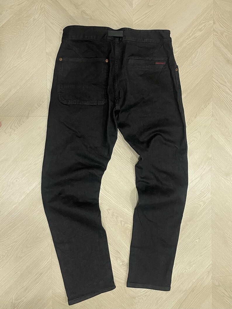 Puede incluir: Pantalones vaqueros negros con un dise&ntilde;o cl&aacute;sico de cinco bolsillos. Los pantalones tienen cierre de bot&oacute;n, trabillas para el cintur&oacute;n y una peque&ntilde;a etiqueta roja con el logotipo en el bolsillo trasero. Los pantalones est&aacute;n colocados sobre una superficie de madera clara.
