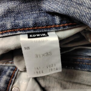 May include: Close-up of blue denim jeans with a white tag. The tag reads "EDWIN" and "SIZE 31x33". The jeans have a button closure and brown stitching. The fabric is faded and worn.