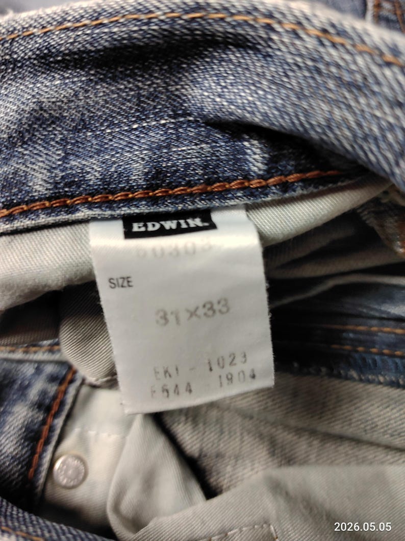 May include: Close-up of blue denim jeans with a white tag. The tag reads "EDWIN" and "SIZE 31x33". The jeans have a button closure and brown stitching. The fabric is faded and worn.