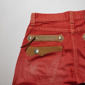 May include: Red denim jeans with brown leather accents. Features include zippered pockets, decorative stitching, and a leather patch with the word "Exclusive". The back pocket has a stitched design. The brand name "Edwin" is visible.