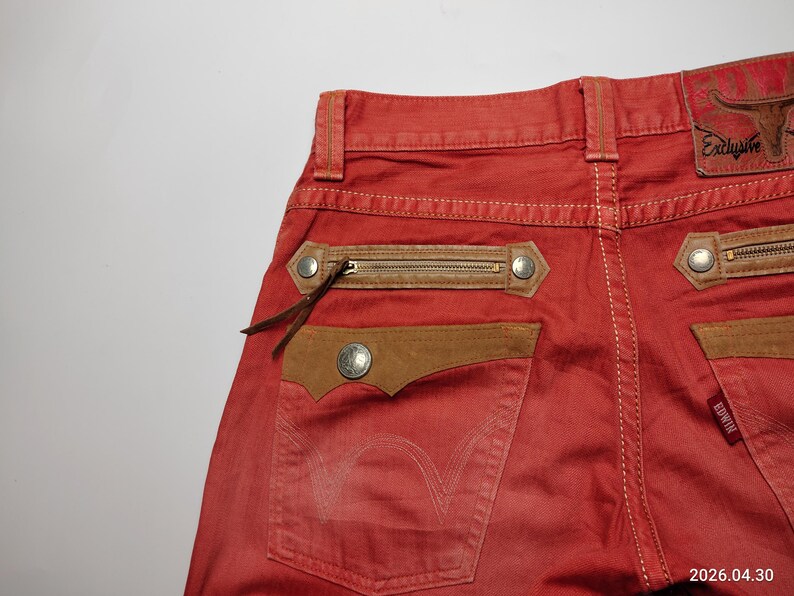 May include: Red denim jeans with brown leather accents. Features include zippered pockets, decorative stitching, and a leather patch with the word "Exclusive". The back pocket has a stitched design. The brand name "Edwin" is visible.