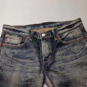 May include: Close-up of blue denim jeans with a button closure and pockets. The jeans have a faded, worn look with visible stitching and rivets. The brand name "EDWIN" is visible on the waistband.