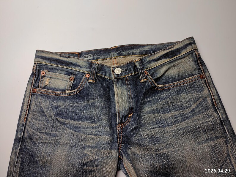 May include: Close-up of blue denim jeans with a button closure and pockets. The jeans have a faded, worn look with visible stitching and rivets. The brand name "EDWIN" is visible on the waistband.