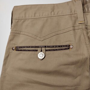 May include: Close-up of khaki-colored pants with a decorative back pocket. The pocket features a dark brown leather trim and a silver button with a floral design. The stitching is visible, and the overall style is casual.