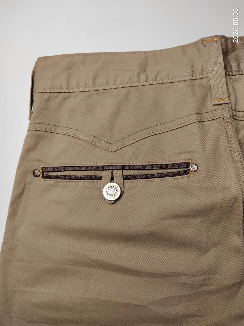 May include: Close-up of khaki-colored pants with a decorative back pocket. The pocket features a dark brown leather trim and a silver button with a floral design. The stitching is visible, and the overall style is casual.