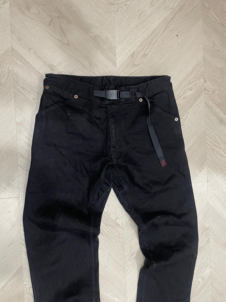 Puede incluir: Pantalones vaqueros negros con cintur&oacute;n negro incorporado. Los pantalones tienen un corte de pierna recta y un peque&ntilde;o logotipo rojo cerca del bolsillo derecho. Est&aacute;n colocados sobre una superficie de vetas de madera clara.