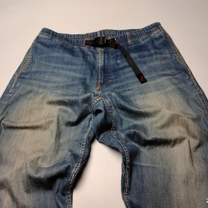 May include: Blue denim trousers with a black belt. The trousers have a faded wash and a relaxed fit. The belt is made of a black fabric and has a black buckle. The trousers are laid flat on a white surface.