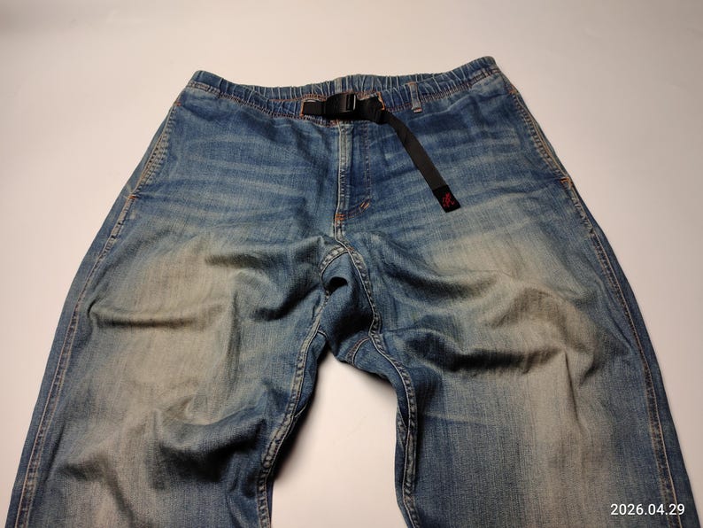 May include: Blue denim trousers with a black belt. The trousers have a faded wash and a relaxed fit. The belt is made of a black fabric and has a black buckle. The trousers are laid flat on a white surface.