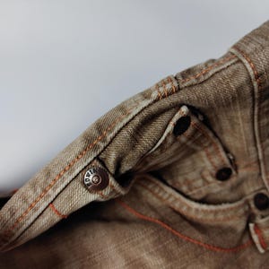 May include: Close-up of brown denim jeans with orange stitching. The jeans feature a metal button with the brand name and additional metal rivets. The fabric has a worn appearance, and the stitching is visible along the seams and pockets.
