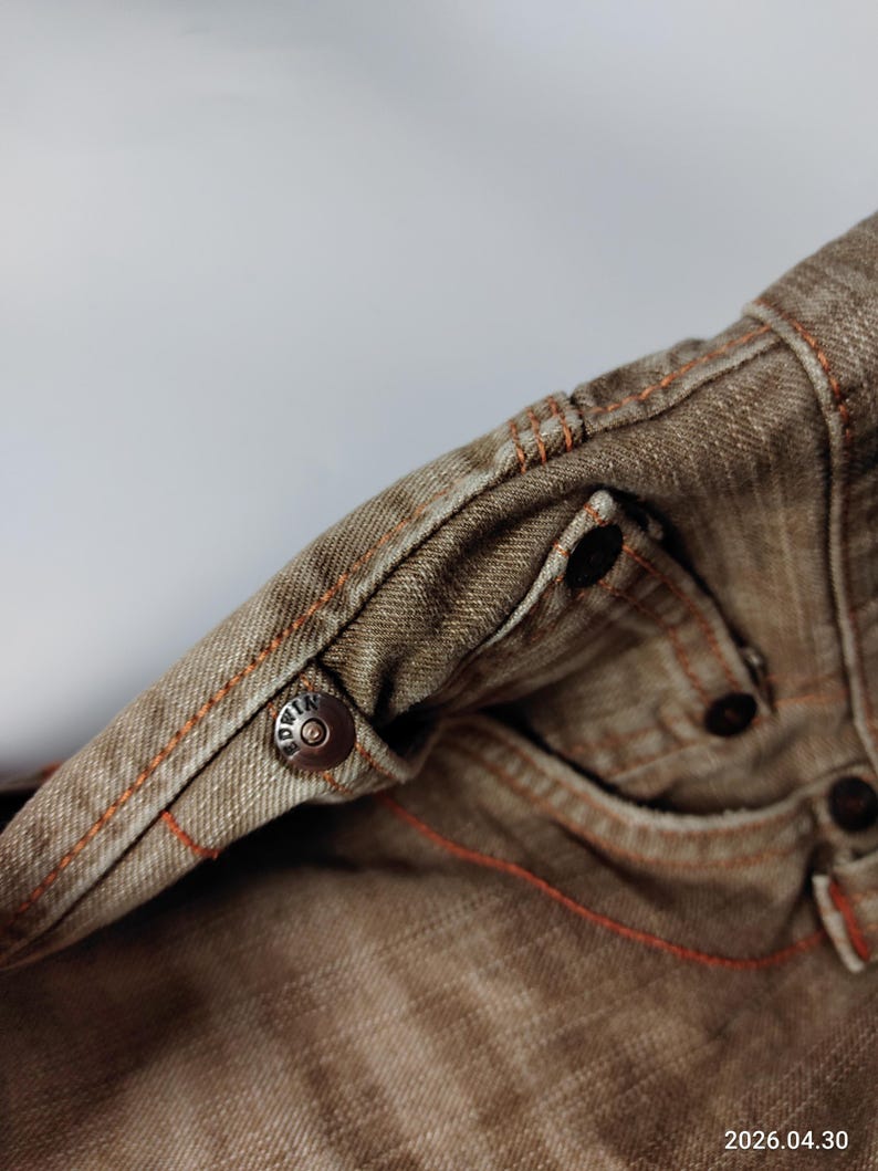 May include: Close-up of brown denim jeans with orange stitching. The jeans feature a metal button with the brand name and additional metal rivets. The fabric has a worn appearance, and the stitching is visible along the seams and pockets.