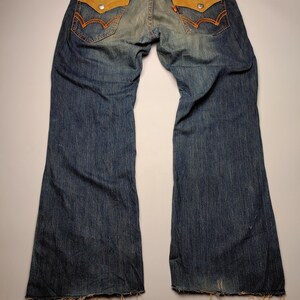 May include: Blue denim jeans with light brown suede-like back pockets and orange stitching. The jeans have a faded wash and a wide leg. The Edwin brand label is visible on the back. The jeans are laid flat on a white surface.